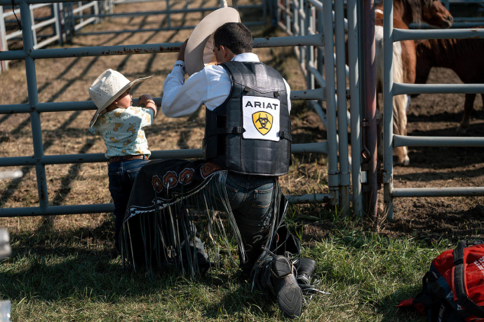 A photograph of man in an Ariat vest and cowboy hat talking to a child at the rodeo